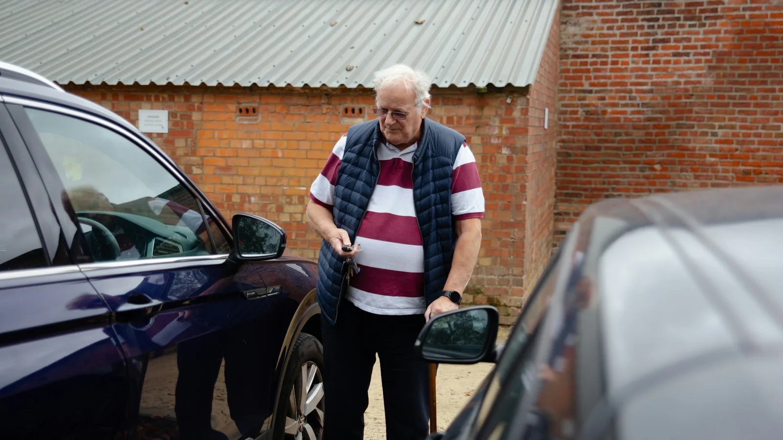 An older man holding car keys while standing between two cars in a gravel driveway