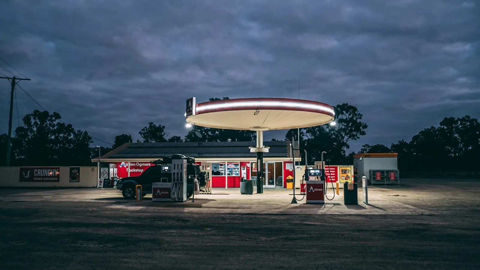 A remote roadhouse lit up at dusk with a vehicle parked at the fuel bowsers