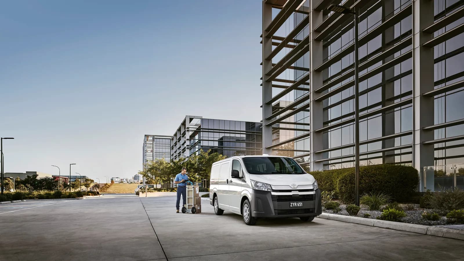 A white Toyota HiAce van parked outside an office building with a parcel trolley alongside