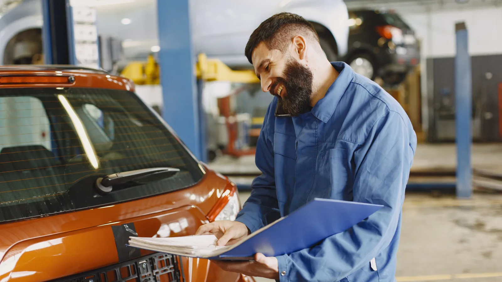 A mechanic in overalls checking paperwork on a clipboard beside a car in a workshop