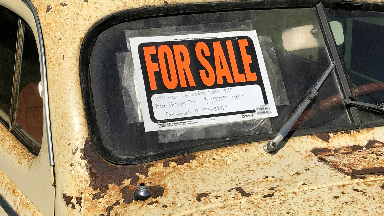 A handwritten for sale sign taped inside the rear window of a rusted old car