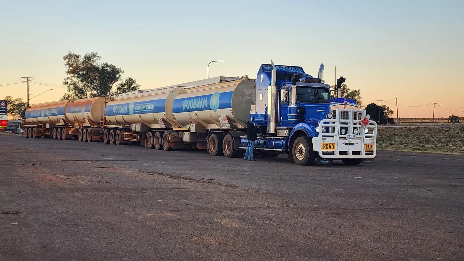 A multi-trailer fuel tanker road train parked at a roadhouse at dusk with its driver standing alongside