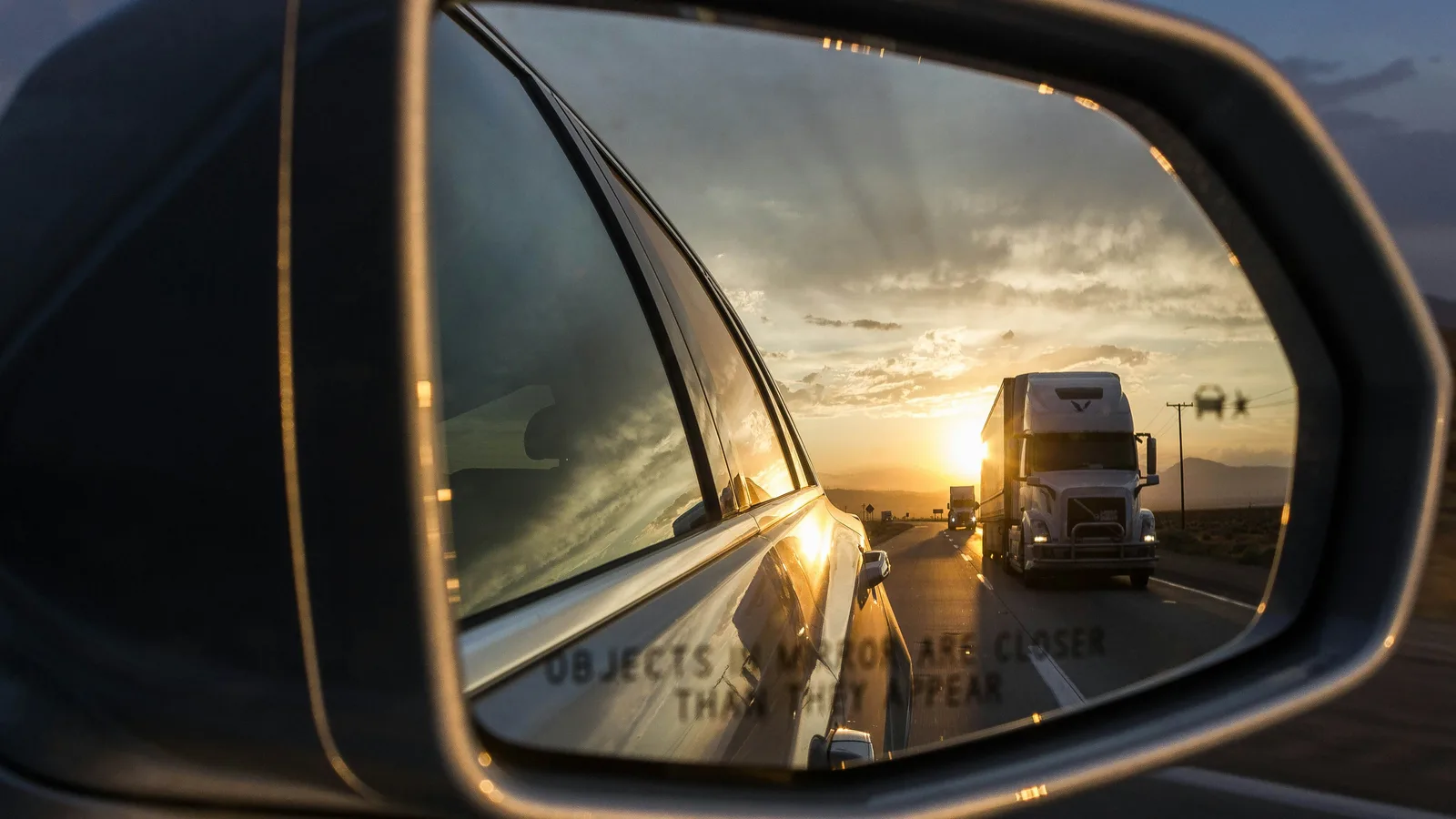 A semi-trailer reflected in a car's wing mirror on a highway at sunset
