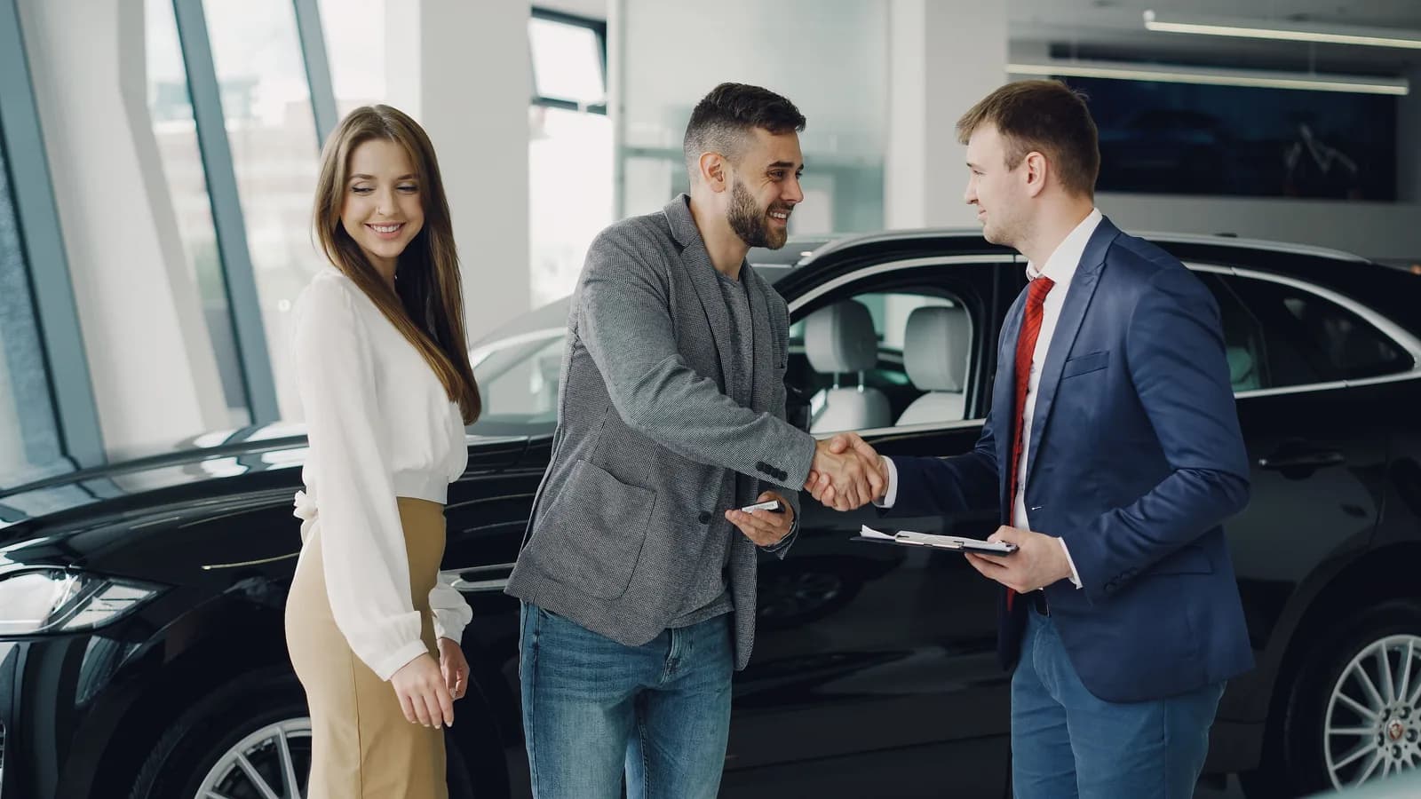 A buyer and a salesperson shaking hands next to a black car in a showroom
