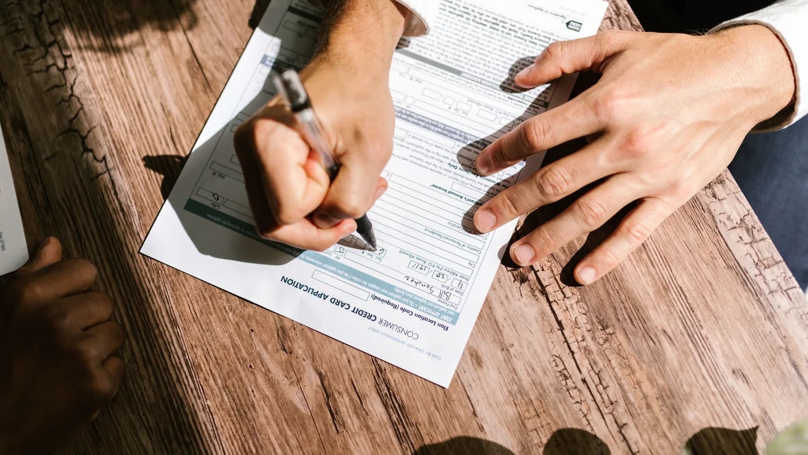 A hand filling in a printed loan application form at a wooden table outdoors