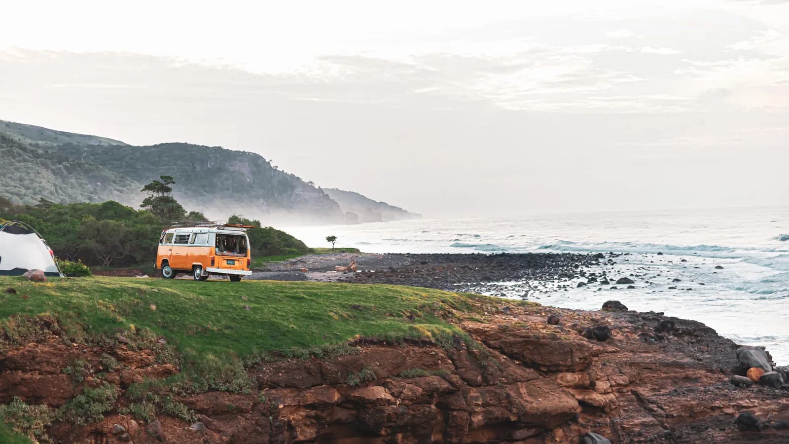 An orange VW Kombi camper parked on a grassy clifftop above the ocean with a tent alongside it