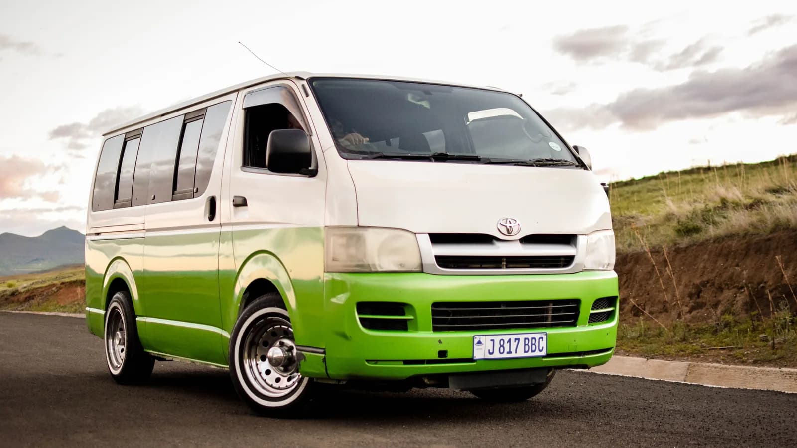 A white and green Toyota HiAce van parked on a rural road, seen from a front three-quarter angle