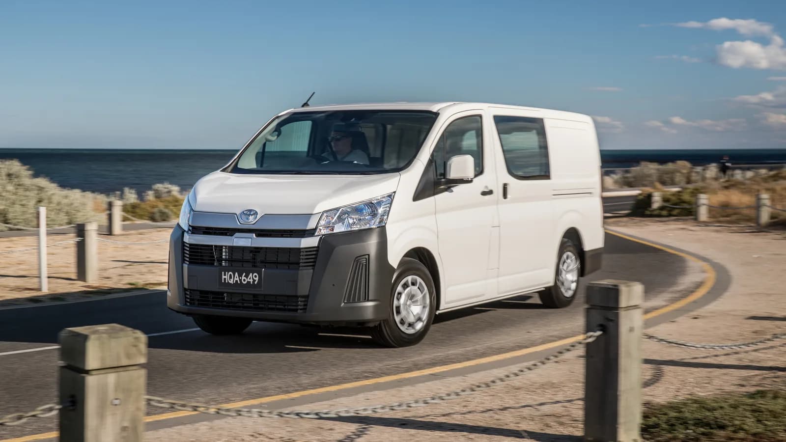 A white Toyota HiAce van driving round a bend beside a beach fence and the sea