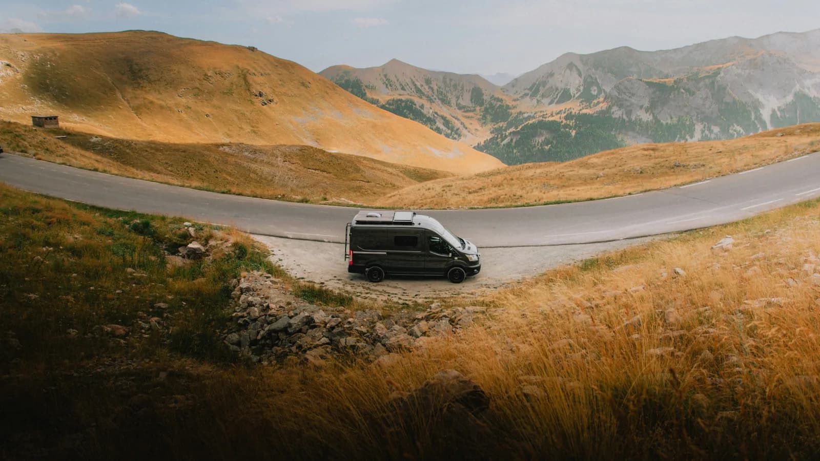 A dark Ford Transit campervan with roof solar panels parked at a pull-off beside a mountain road, side-front view