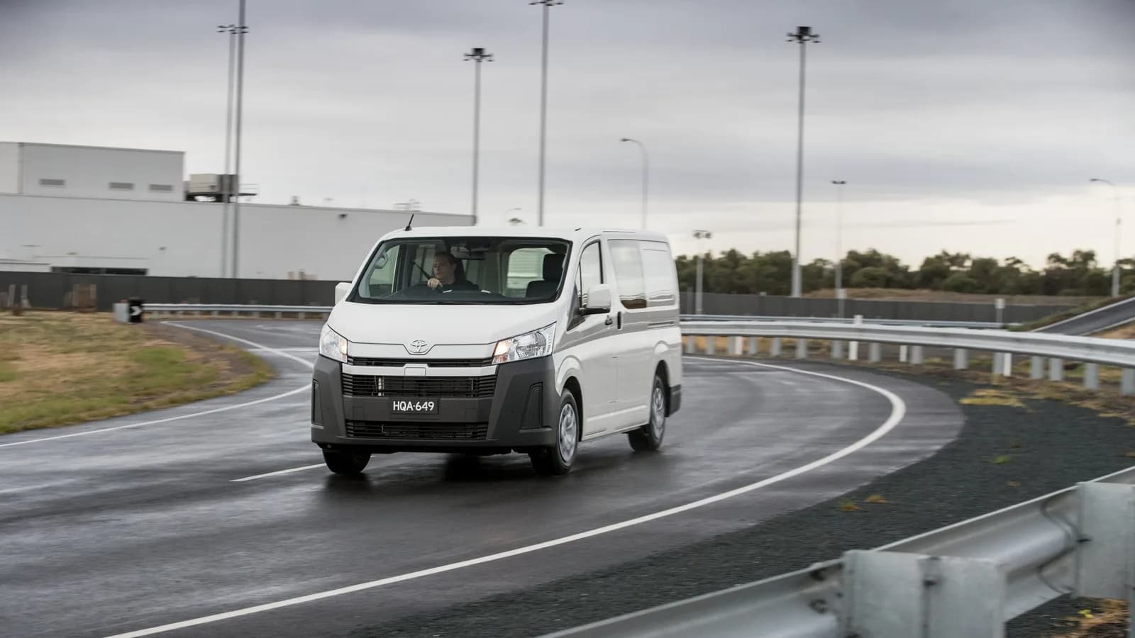 A white HiAce van cornering on a wet road with guardrails, front three-quarter view