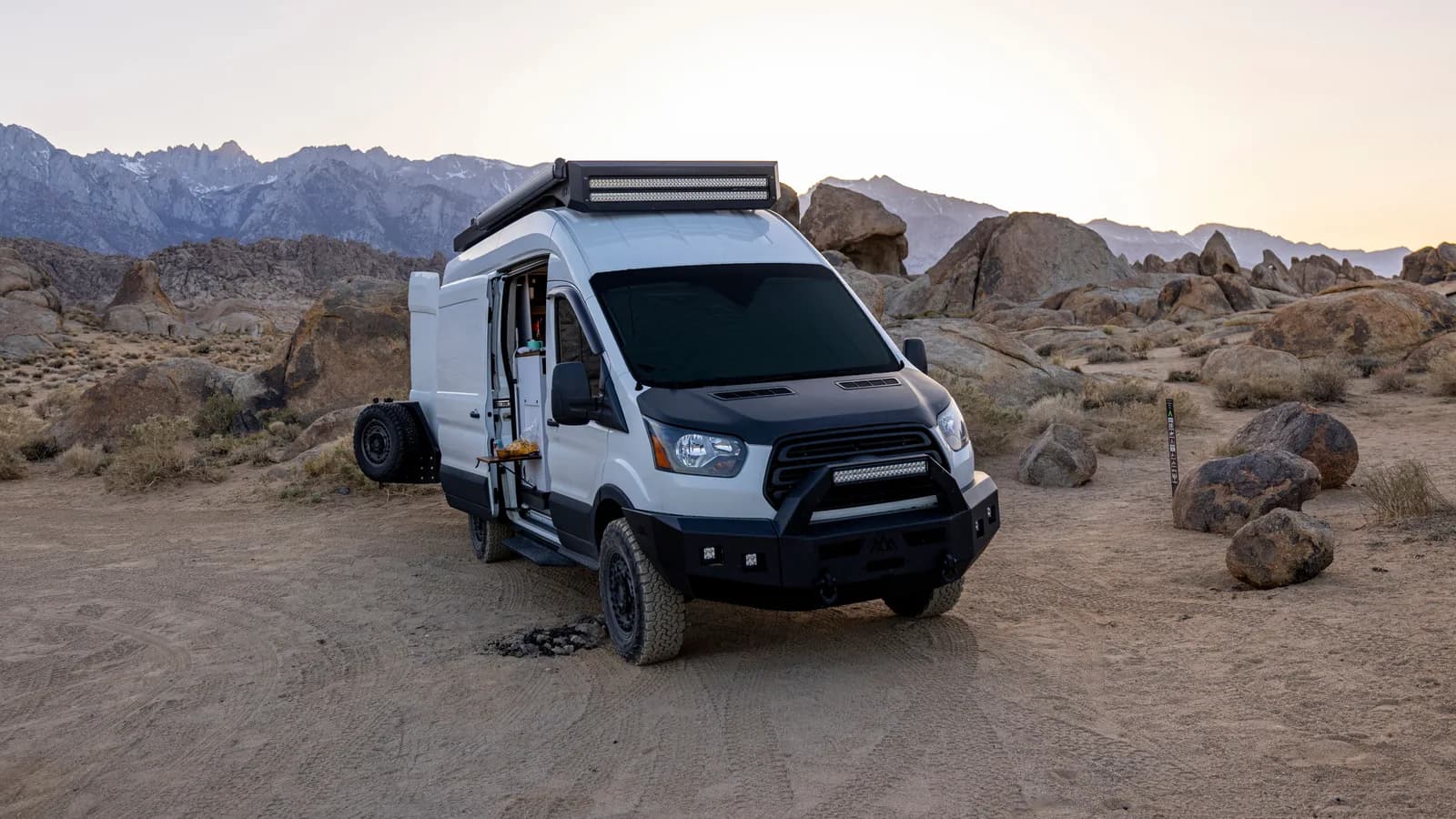 A white Ford Transit camper with bull bar and roof light bar parked at a desert campsite among boulders, three-quarter front view, side door open