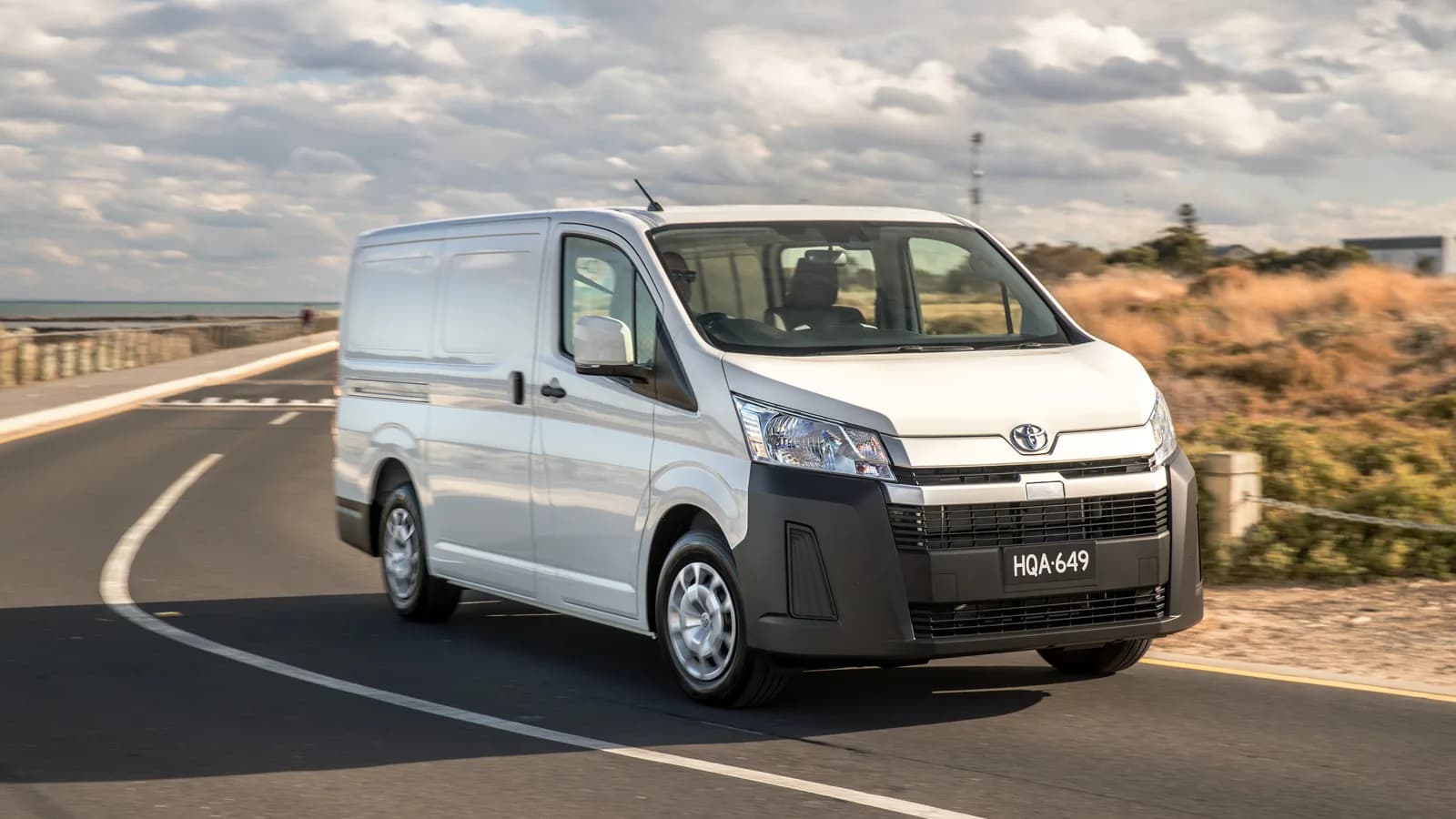 A white Toyota HiAce van driving on a coastal road, three-quarter front view, with dunes and sea behind