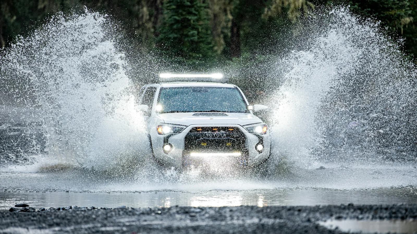 A white four-wheel drive pushing a bow wave through a flooded road crossing
