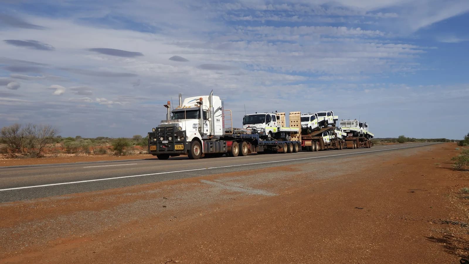 A road train hauling several loaded flatbed trailers along a red dirt outback highway