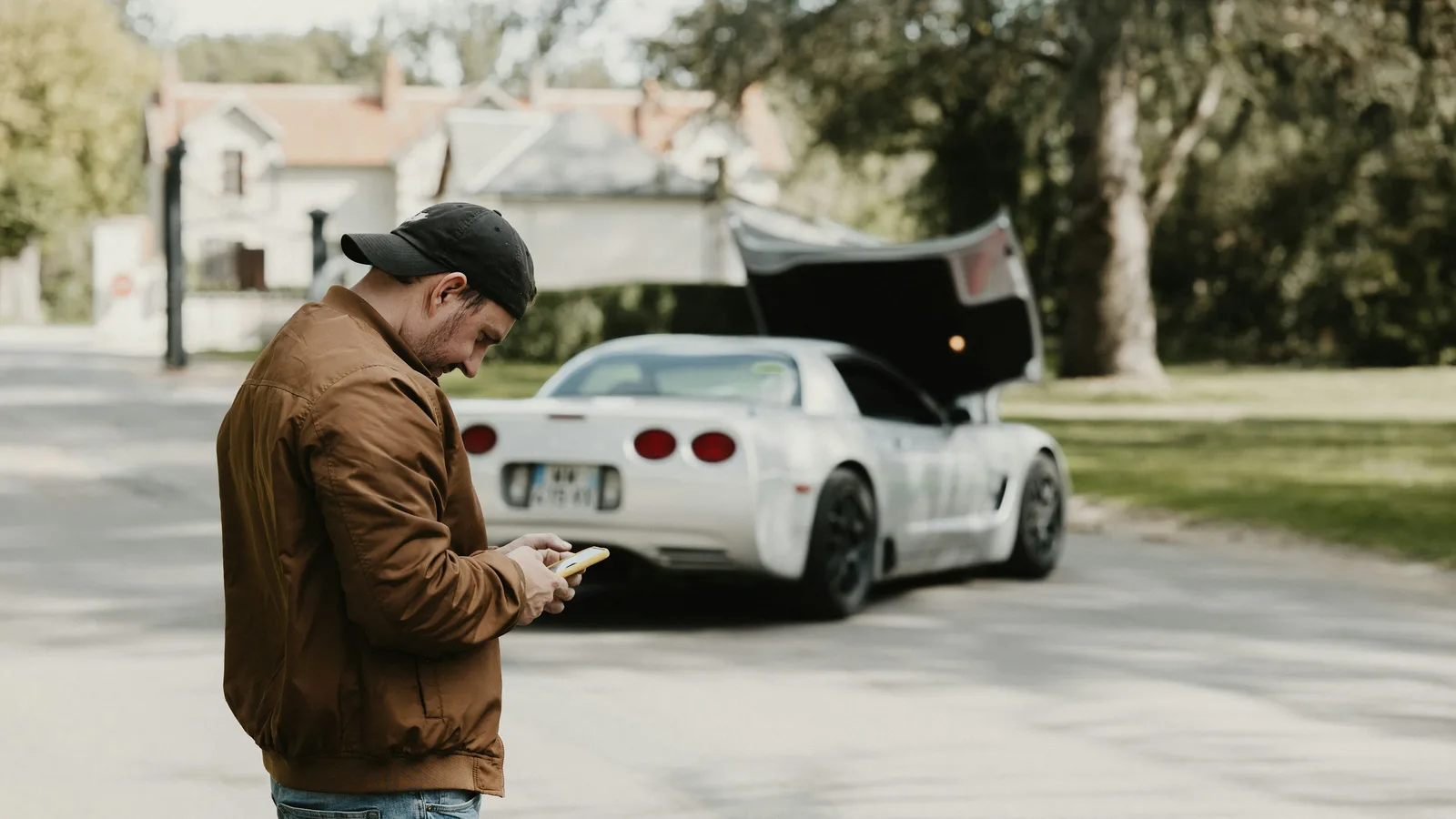 A man standing beside a parked car with the bonnet raised, looking at his phone